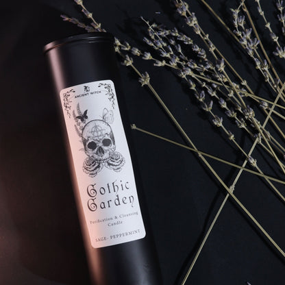 Bougie Gothic Garden noire avec étiquette crâne et roses, parfum sauge-menthe, posée près de branches de lavande.