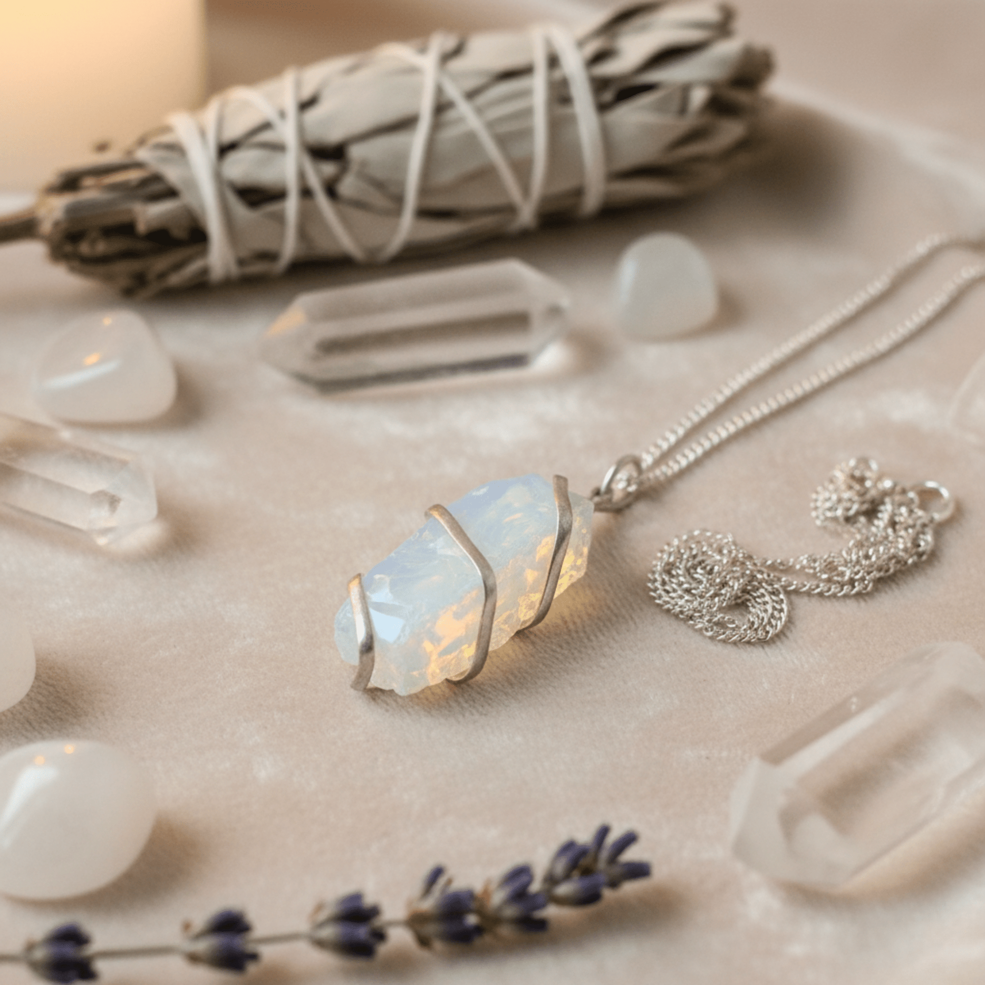 Pendentif opalite brute en fil argenté présenté avec cristaux de roche et sauge blanche sur fond velours clair.