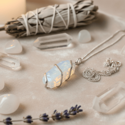 Pendentif opalite brute en fil argenté présenté avec cristaux de roche et sauge blanche sur fond velours clair.