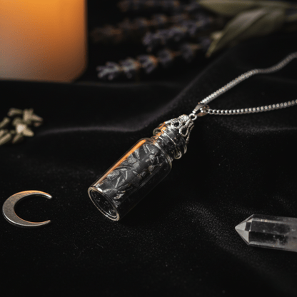 Fiole en verre remplie d’onyx noir sur une chaîne argentée, posée sur tissu noir rituel.