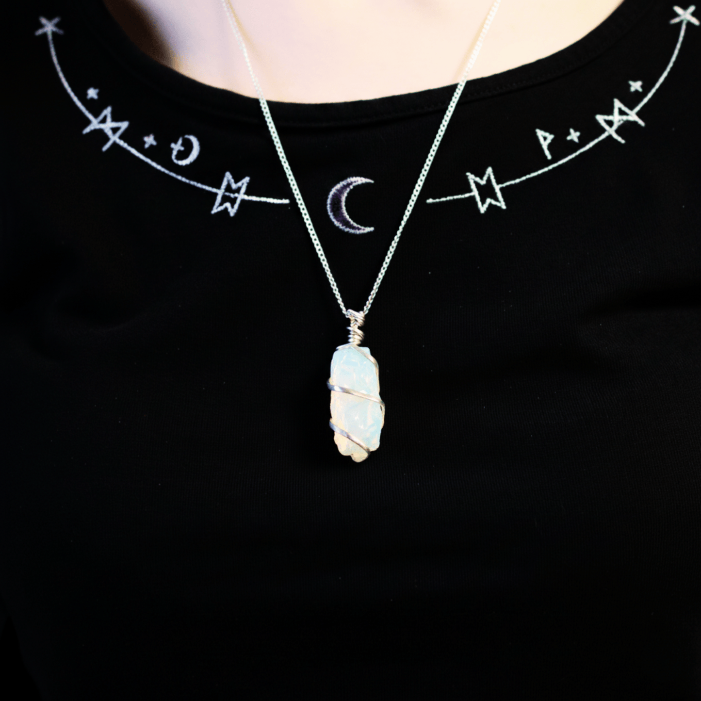 Collier en opalite brute porté autour du cou, pendentif lumineux mis en valeur sur un haut noir décoré de symboles mystiques.