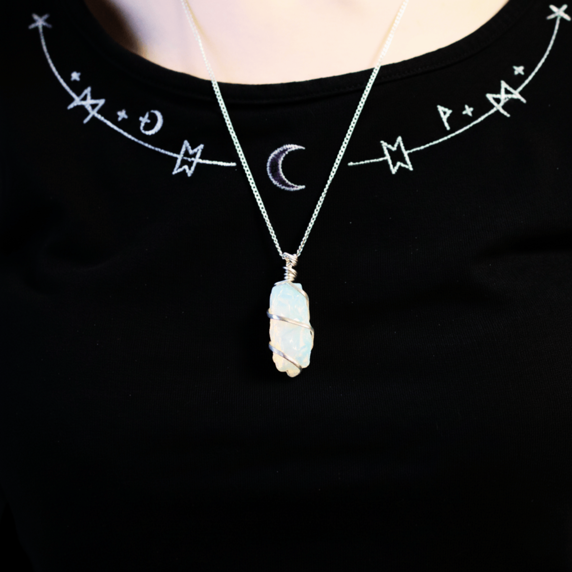 Collier en opalite brute porté autour du cou, pendentif lumineux mis en valeur sur un haut noir décoré de symboles mystiques.