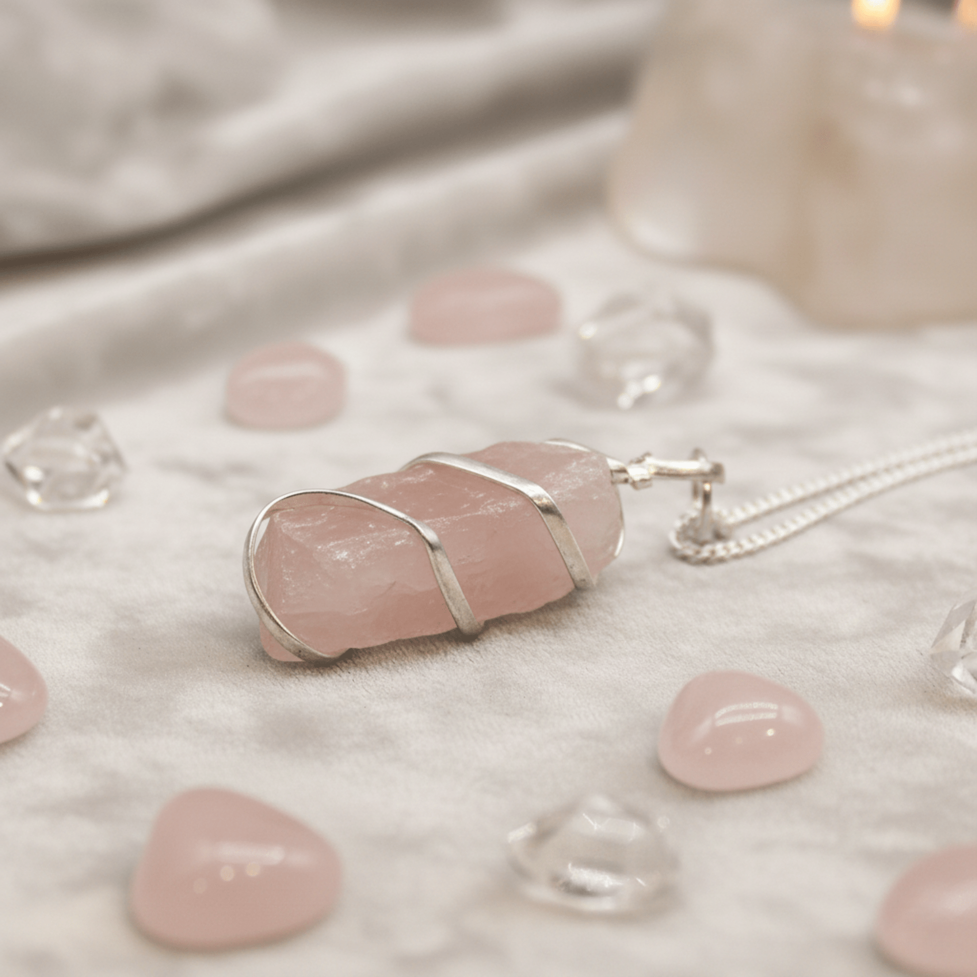 Pendentif quartz rose brut entouré de fil argenté, posé avec pierres polies et cristaux sur tissu clair.