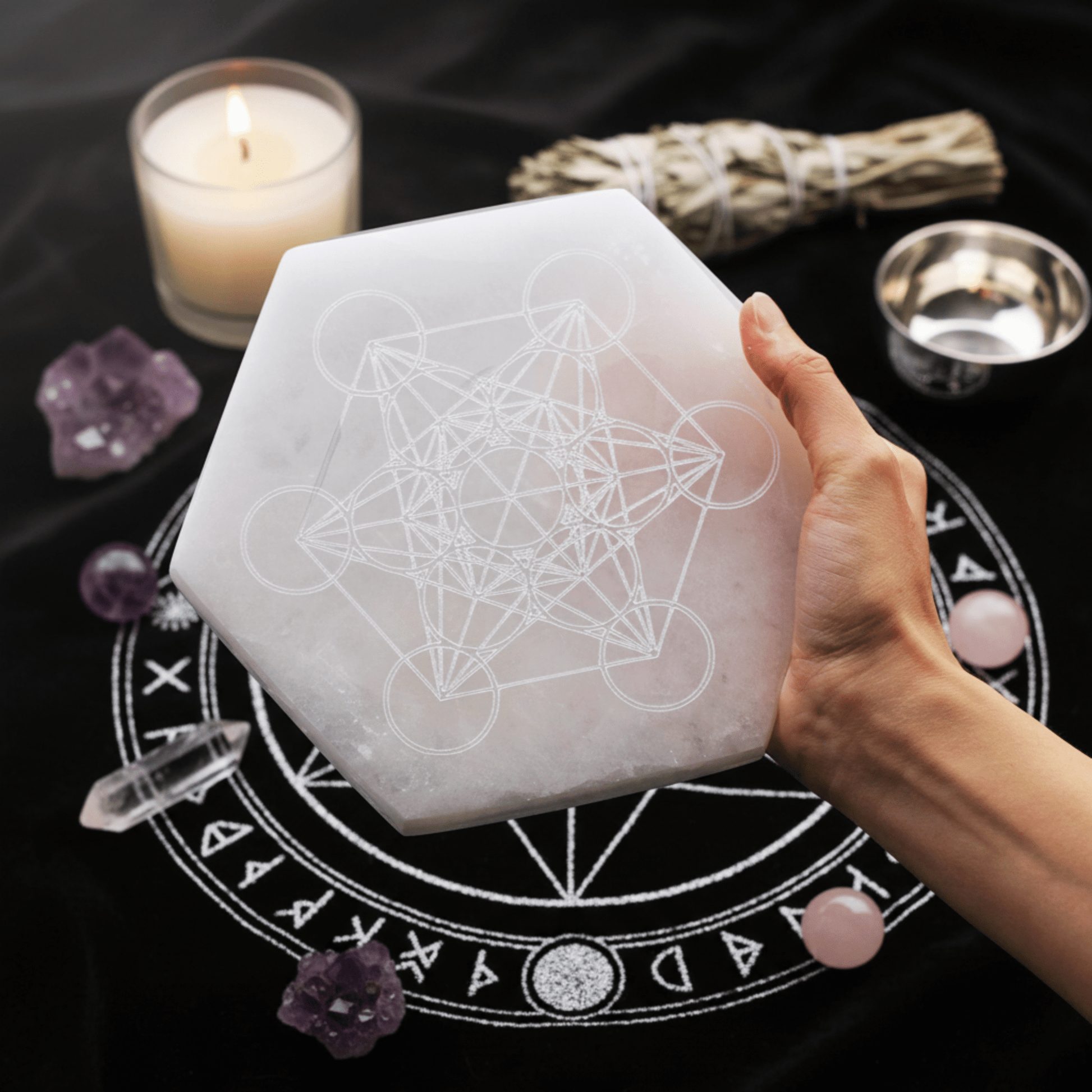 Grande plaque hexagonale en sélénite gravée du Cube de Métatron, tenue en main, avec autel rituel en arrière-plan.